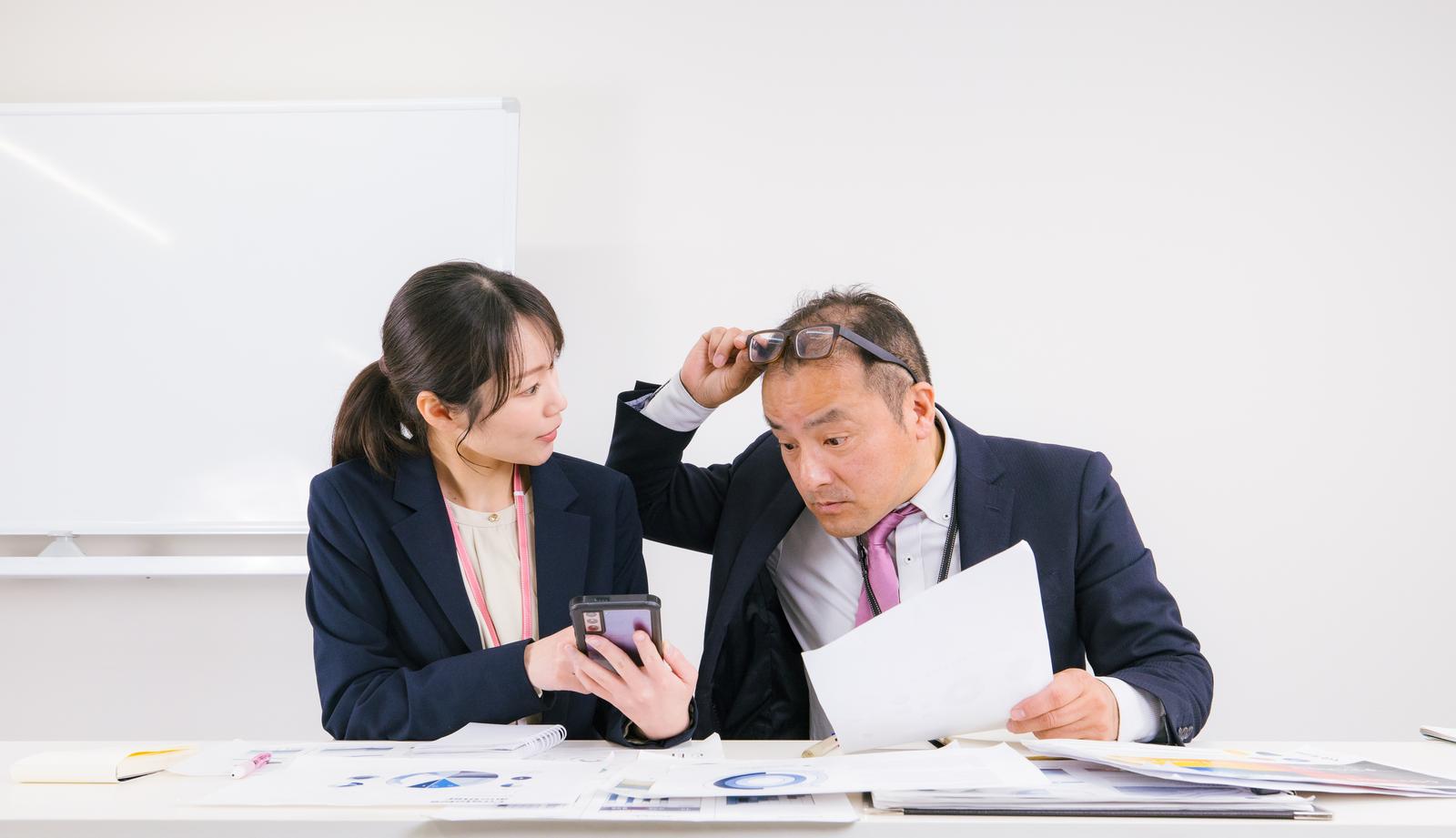 オフィスでスマートフォンと書類を確認し、意見交換する男女の会社員［モデル：高木桂一 SAKI］