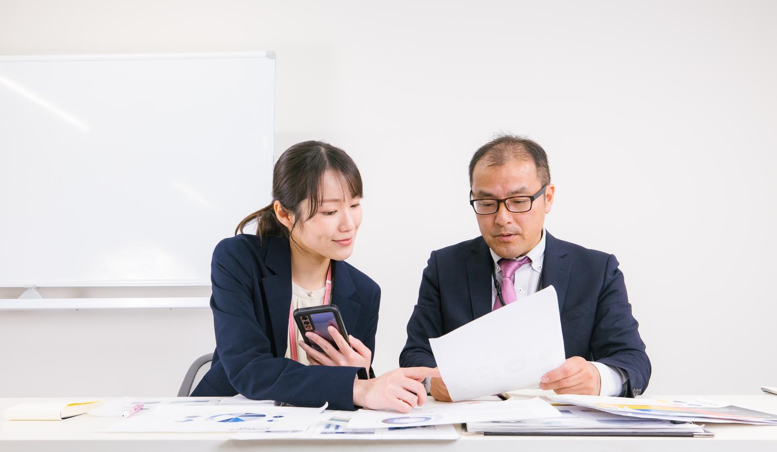 書類とスマートフォンを見ながら打ち合わせする男女の会社員［モデル：高木桂一 SAKI］