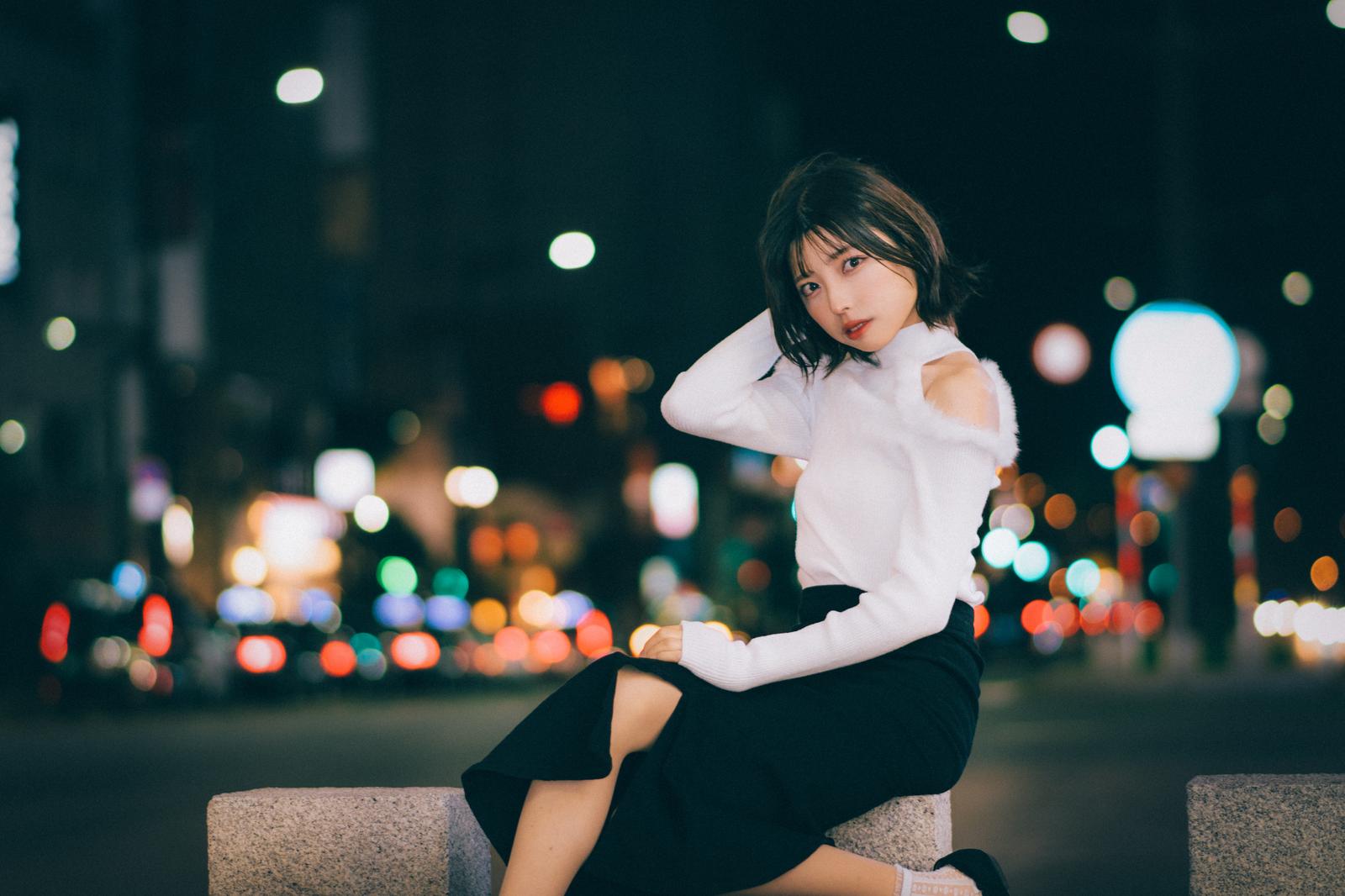 夜の繁華街のネオンが背景に光る中、ベンチに座ってポーズする白シャツ姿の女性［モデル：高尾実生］