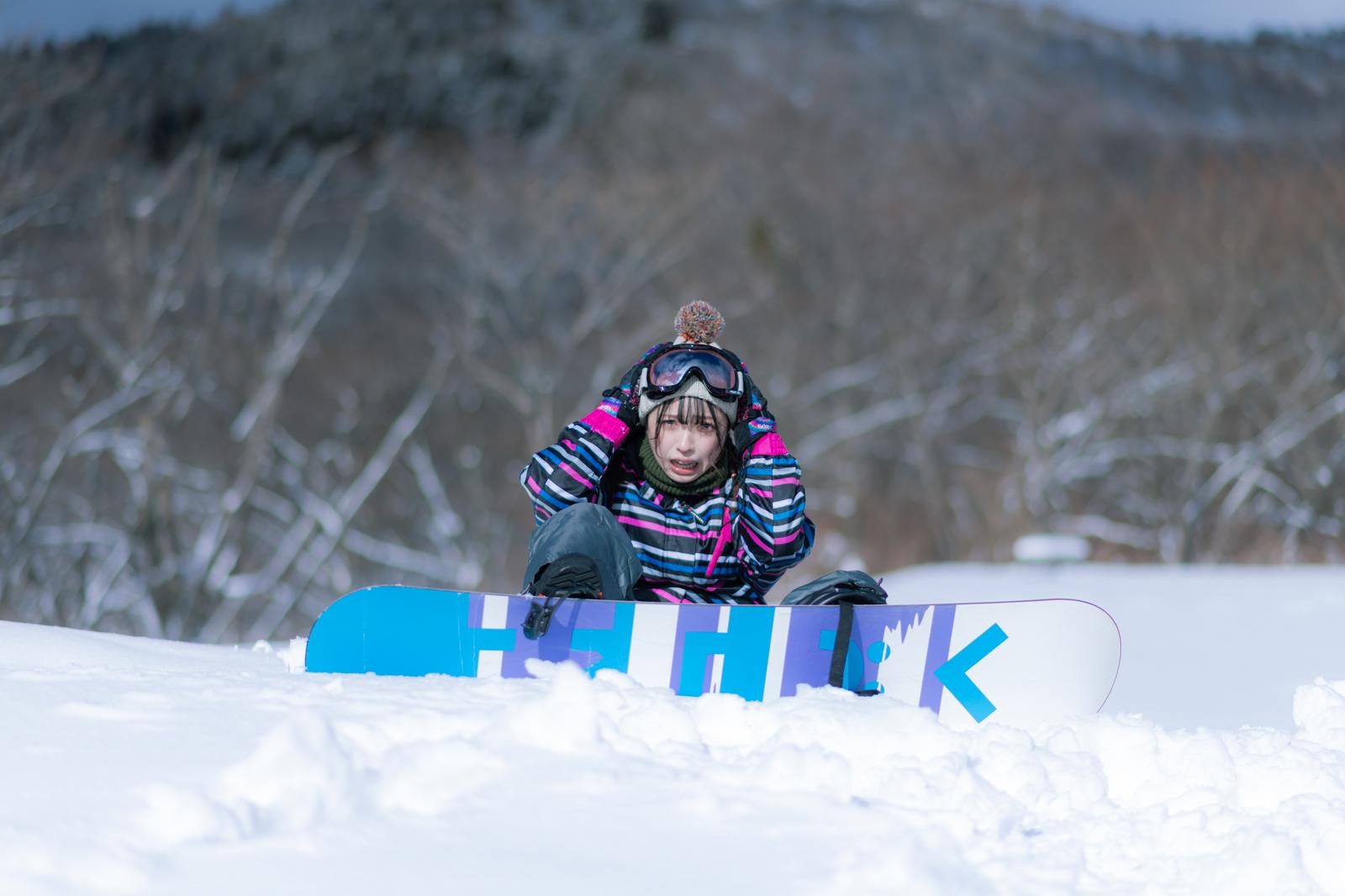 雪山でスノーボードを持ったまま転倒した、カラフルなウェアの女性［モデル：高尾実生］