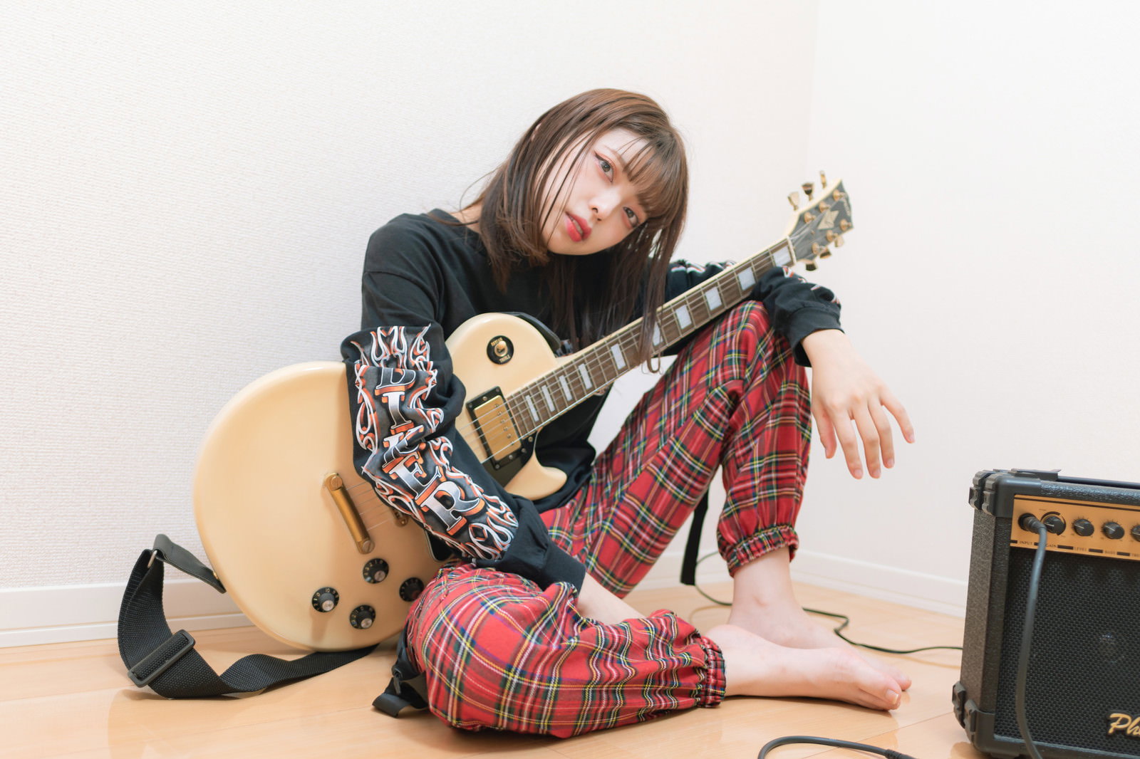 ロックTシャツと赤チェック柄パンツを着た女性がアコースティックギターを膝に抱えて座っている［モデル：高尾実生］
