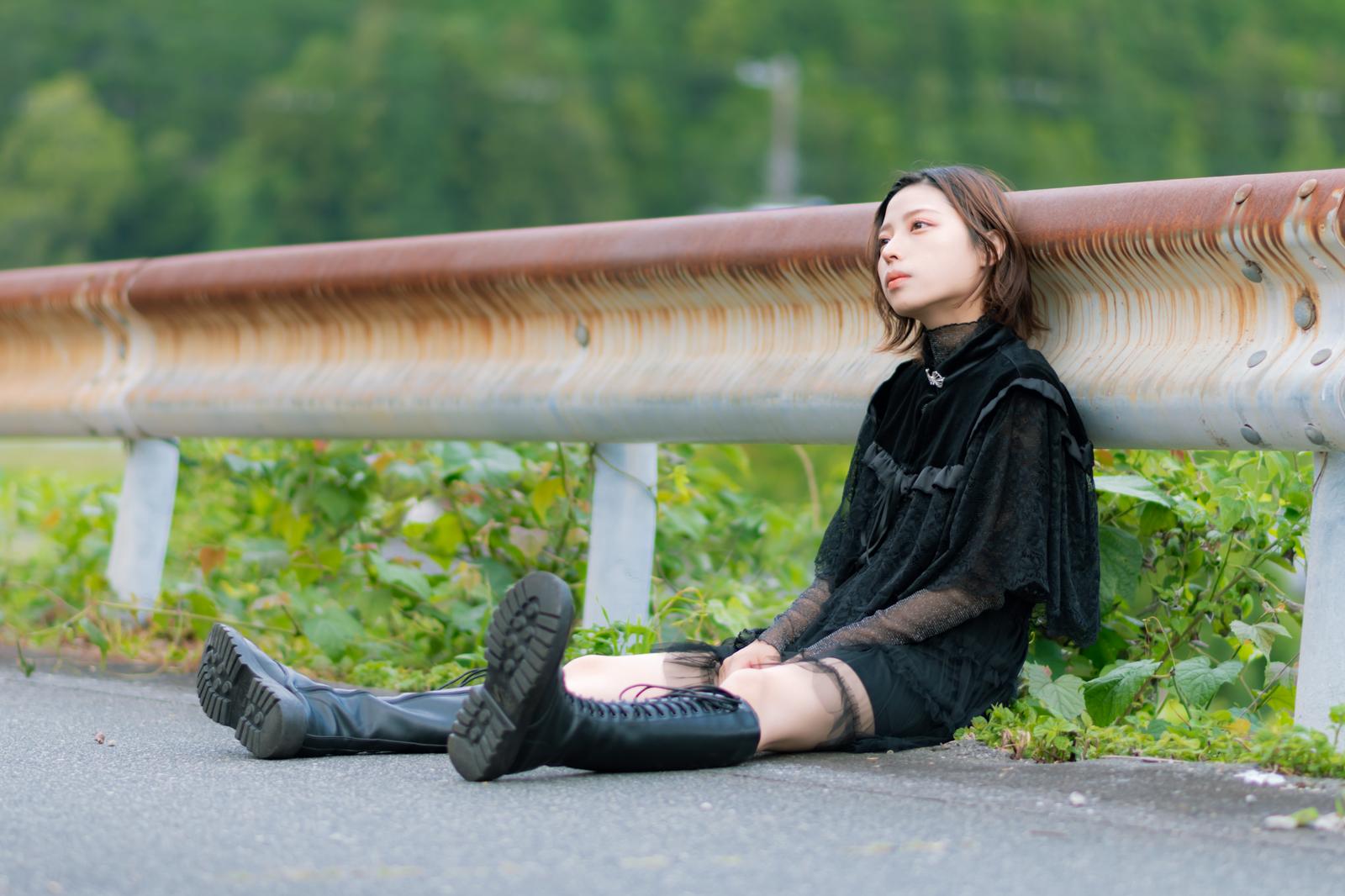 ガードレールに寄りかかって座り込む黒い服の女性のポートレート［モデル：高尾実生］