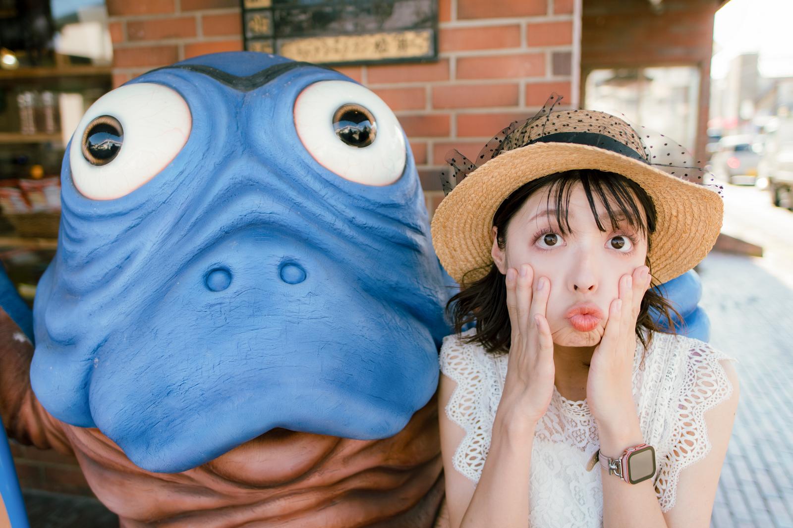 妖怪の海坊主の造形物の横で驚いた表情をする藁帽子の女性［モデル：高尾実生］