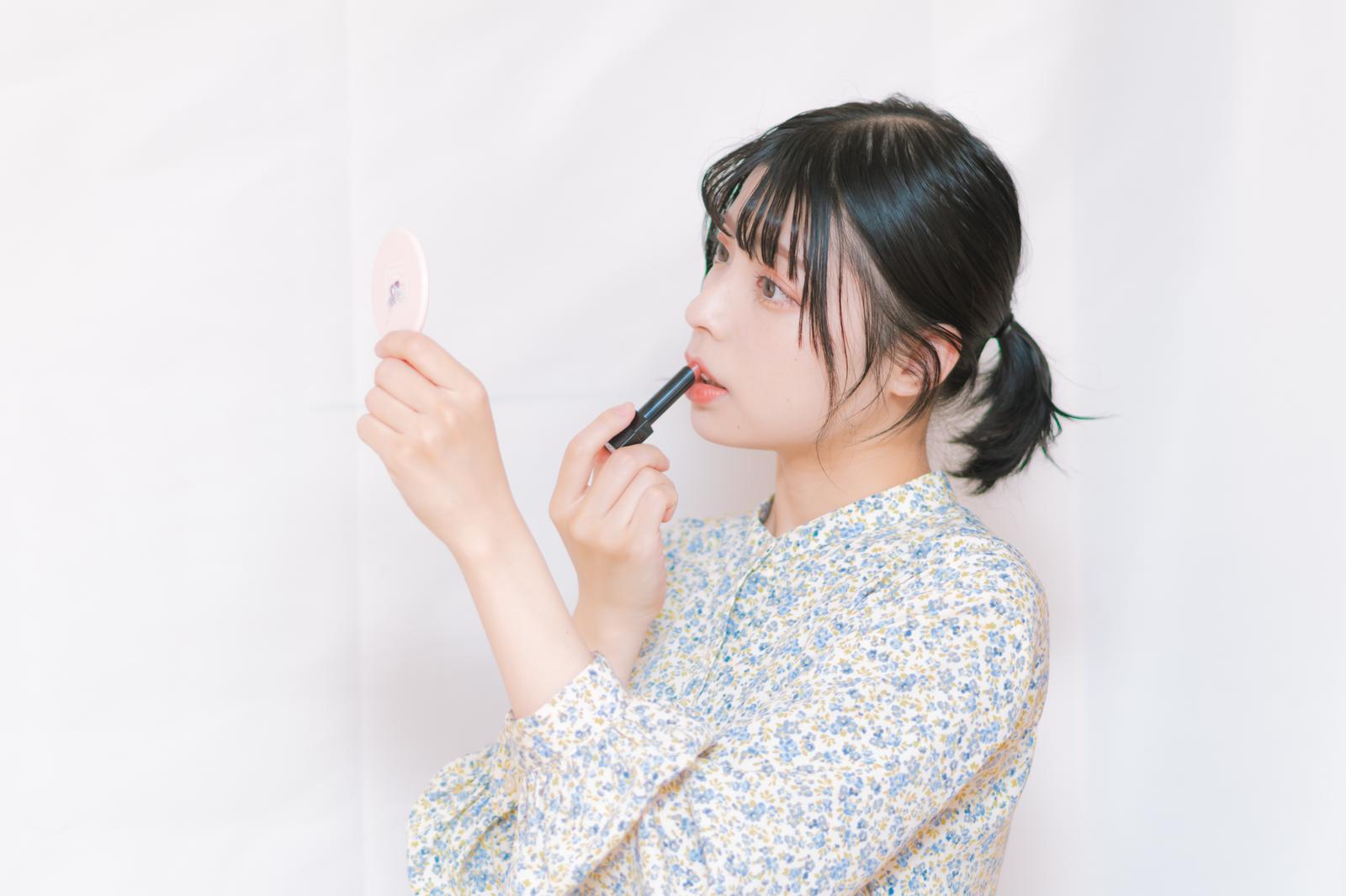 小さな鏡を見ながら黒いリップブラシで口紅を塗る女性［モデル：高尾実生］