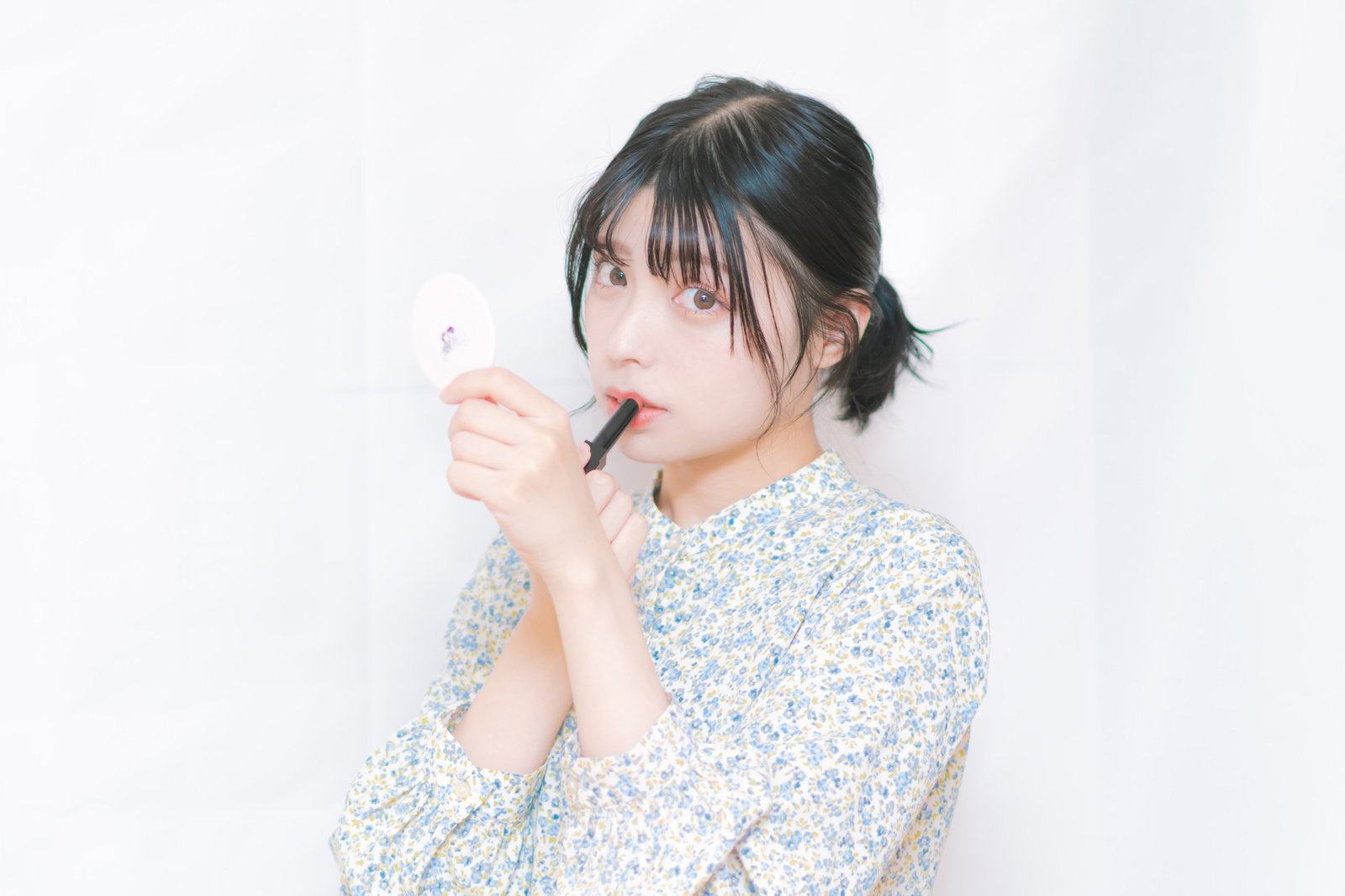 小花柄シャツを着た女性がリップを手に取り鏡を見ながら確認している［モデル：高尾実生］