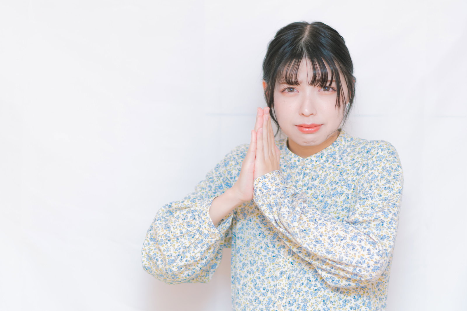 両手を合わせてお願いする表情の女性が白い背景の前に立っている［モデル：高尾実生］