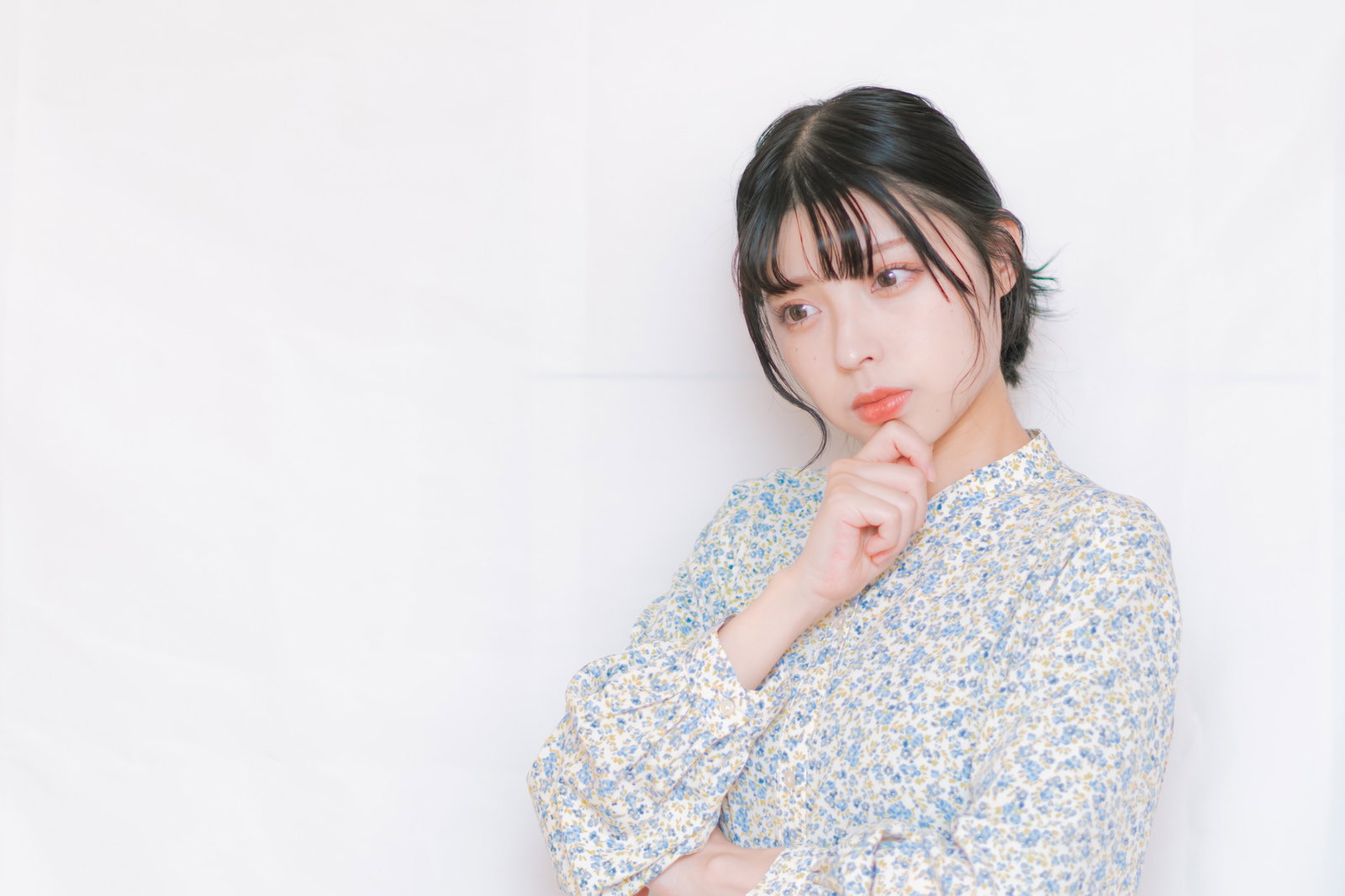 首を傾げて俯瞰し、指を頬に当てて考える青い花柄シャツの女性［モデル：高尾実生］