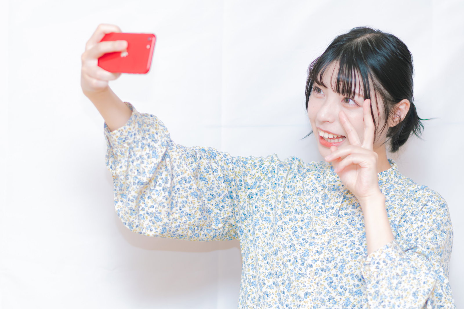 赤いスマホを持ちながらピースをして笑う女性［モデル：高尾実生］