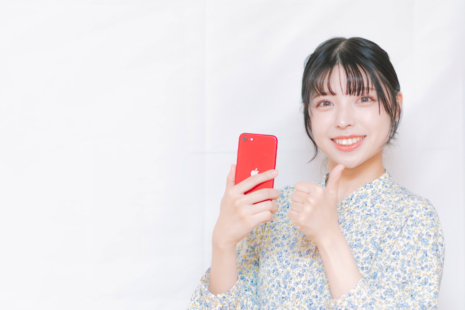 赤いiPhoneを持ち、左手でOKサインをする笑顔の女性。白い背景に黄色と白の花柄シャツ［モデル：高尾実生］