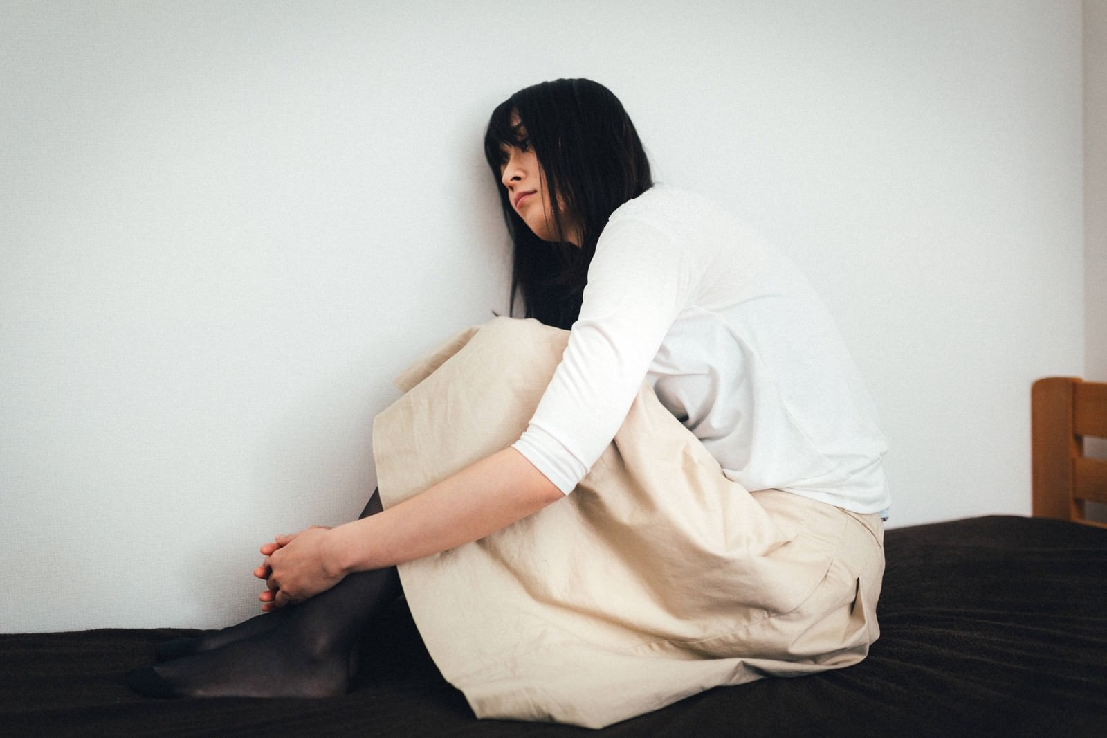 白シャツの女性がベッドで体育座りし壁にもたれる［モデル：たけべともこ］