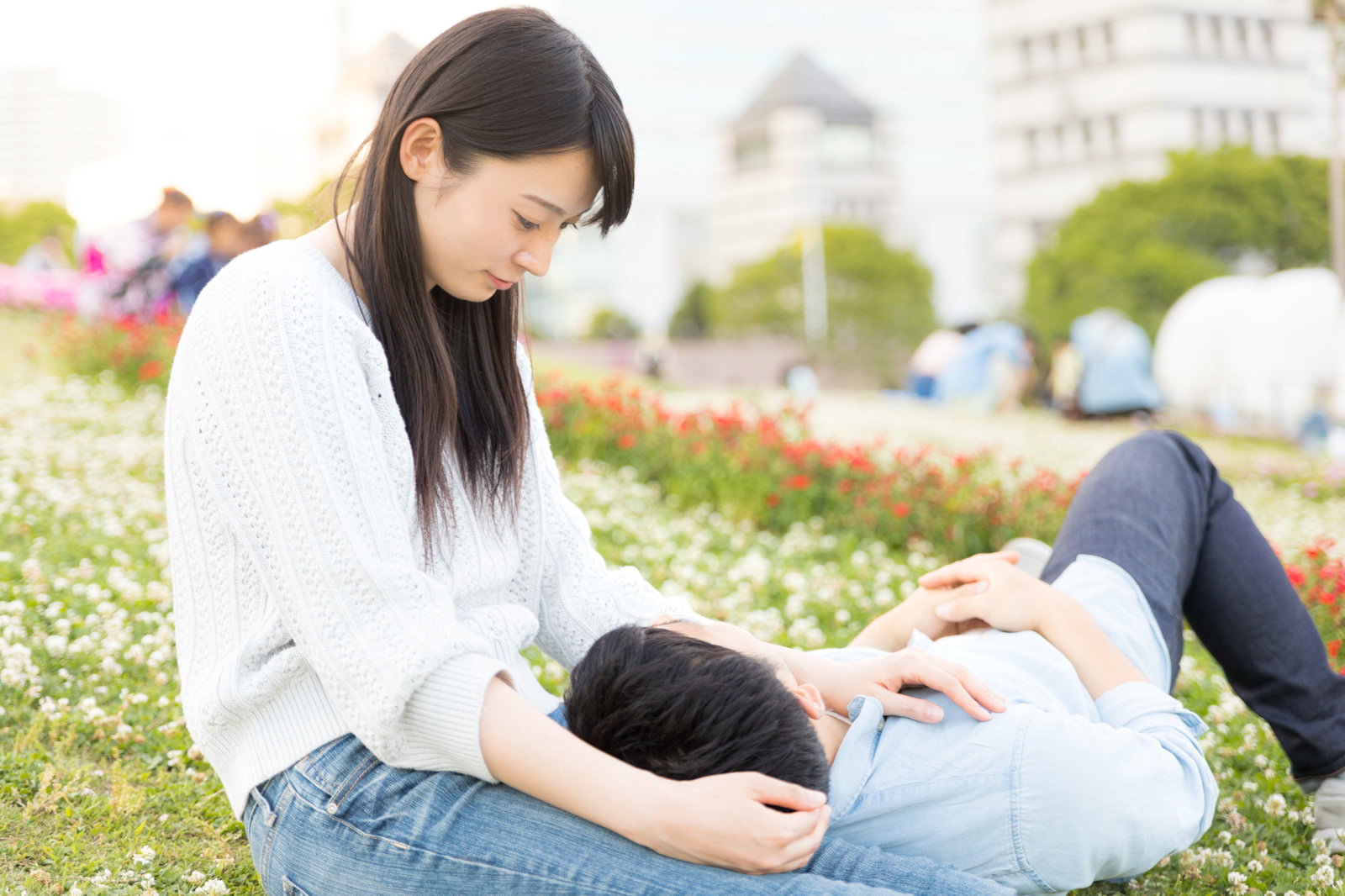 女性が屋外の芝生上で男性の頭を膝に乗せている。昼間の花壇背景［モデル：たけべともこ］