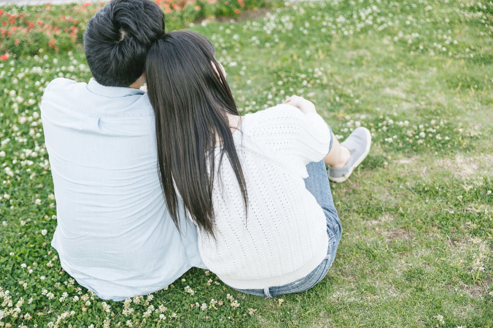 芝生の上に座り、女性が男性の肩にもたれかかっている恋人の後ろ姿［モデル：たけべともこ］