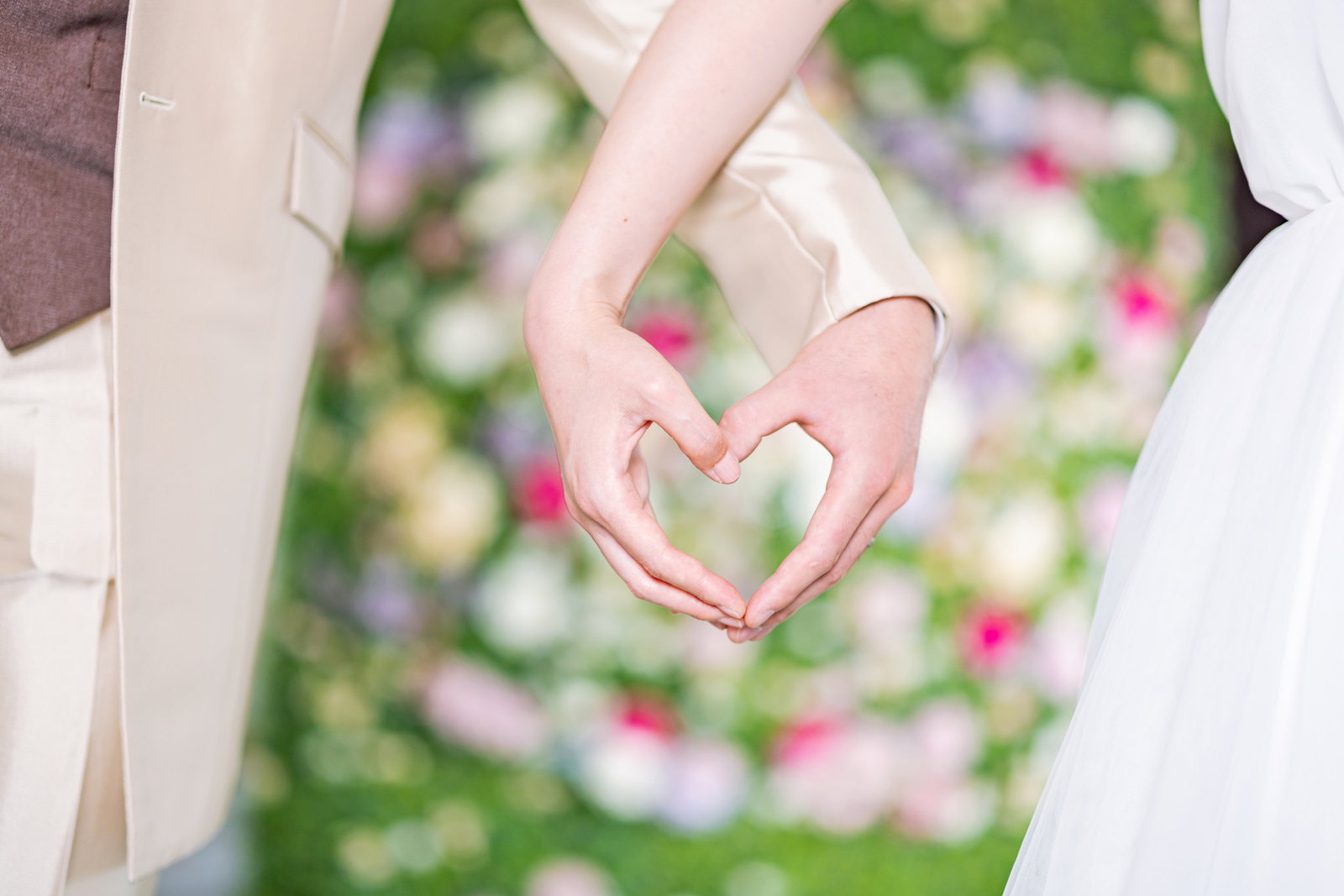 新郎新婦の手で作ったハート型のポーズ、背景に緑とピンクの花［モデル：たけべともこ］