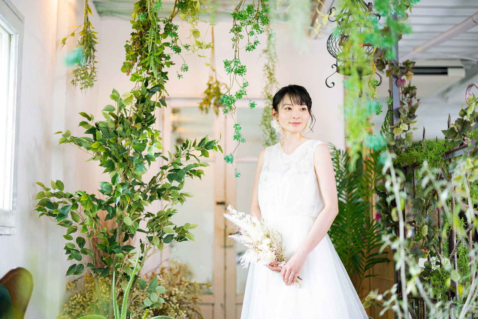 観葉植物に囲まれた明るい室内で白いウェディングドレスを着て微笑む女性［モデル：たけべともこ］