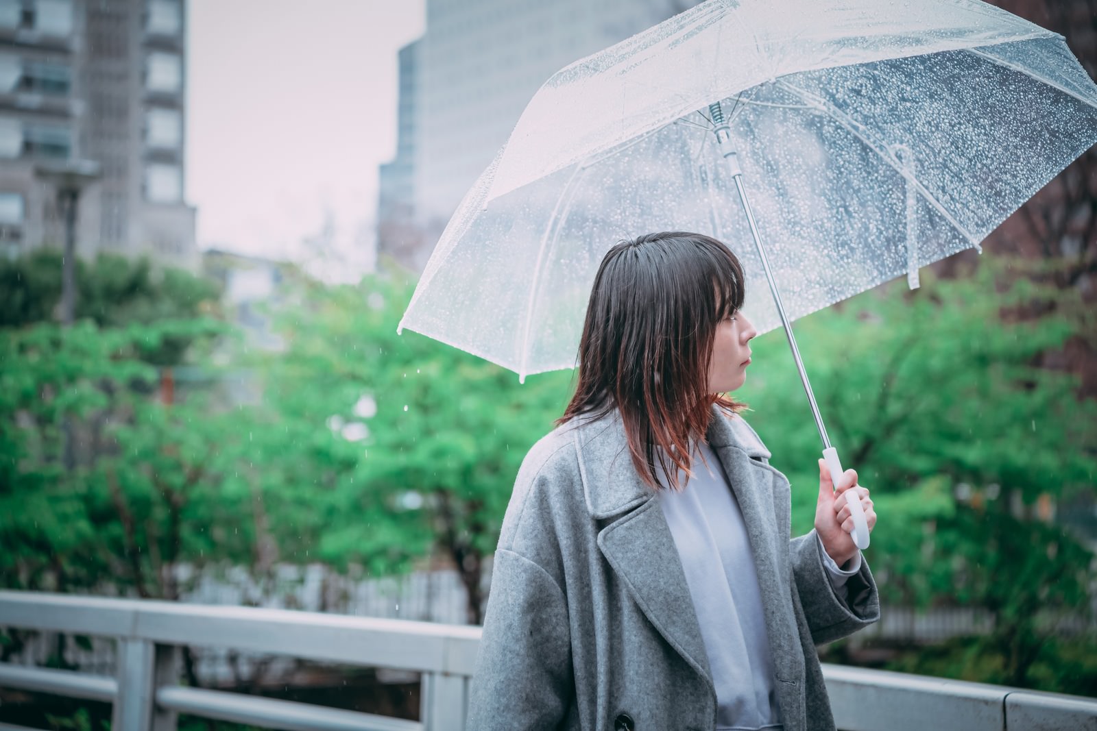 ビニール傘をさしてグレーのコートを着た女性が雨の街を歩く様子［モデル：たけべともこ］
