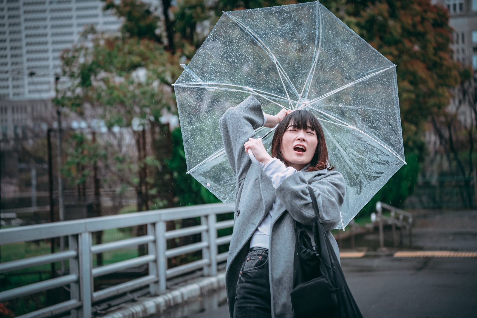 透明なビニール傘を持ち、風に飛ばされそうになる女性の雨の日の写真［モデル：たけべともこ］