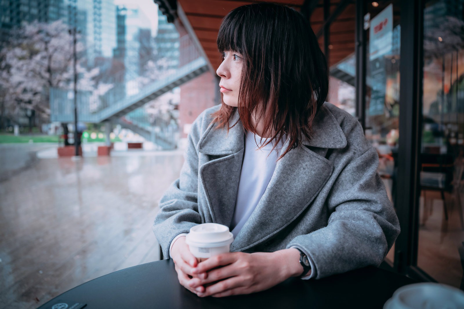 テラス席でコーヒーを持ちながら雨を眺める憂鬱そうな女性［モデル：たけべともこ］
