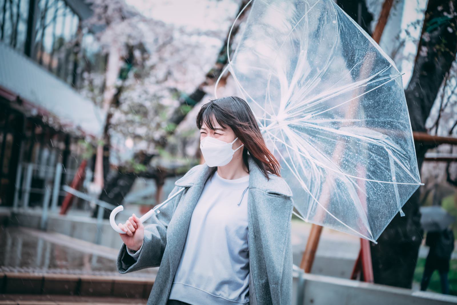 強風で傘が裏返った白いマスク姿の女性がグレーのコート姿で立っている［モデル：たけべともこ］