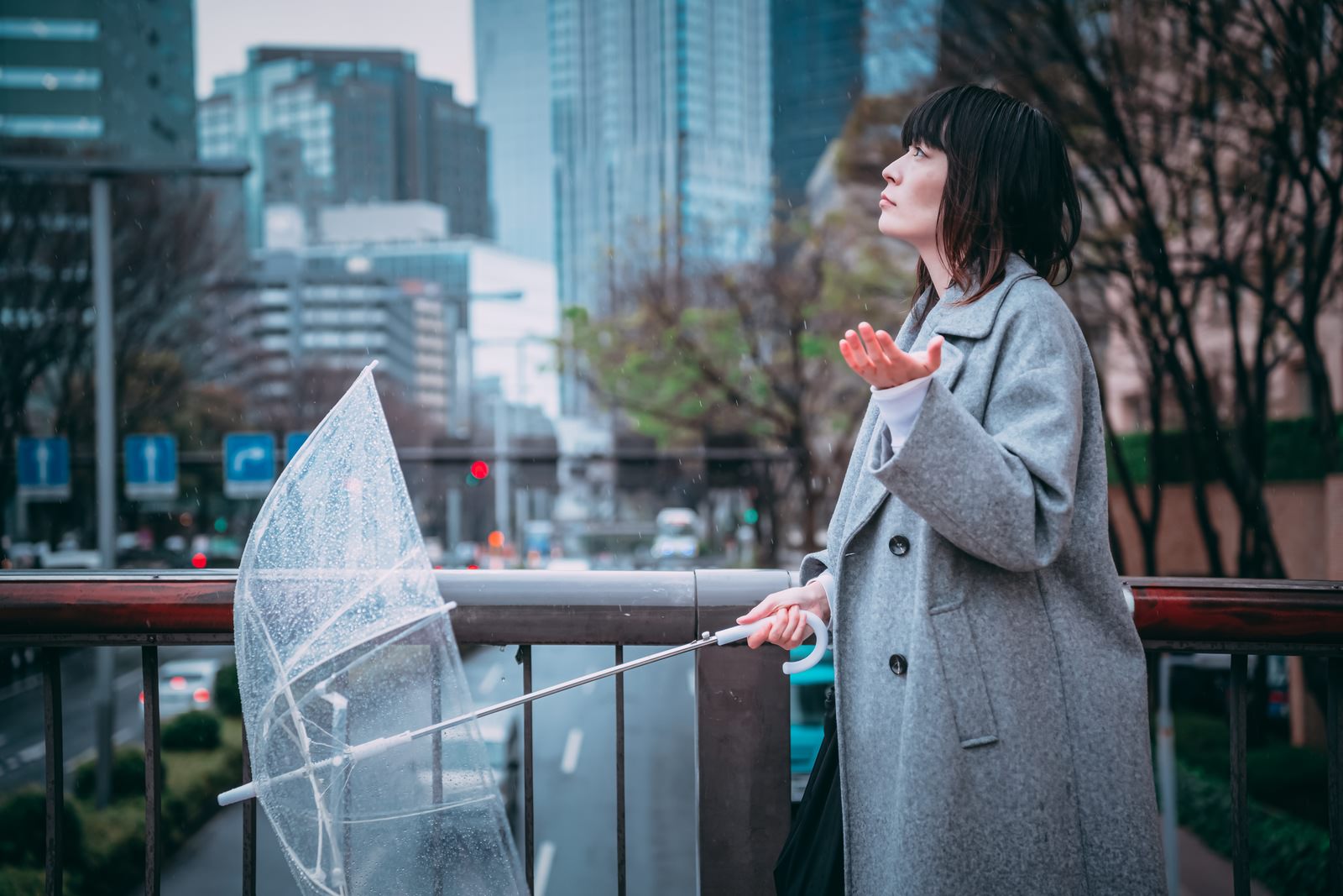 グレーのコートを着た女性が透明なビニール傘を持って雨を確認している［モデル：たけべともこ］