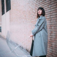 レンガの壁際でビニール傘を持ちながら雨宿りする女性の写真
