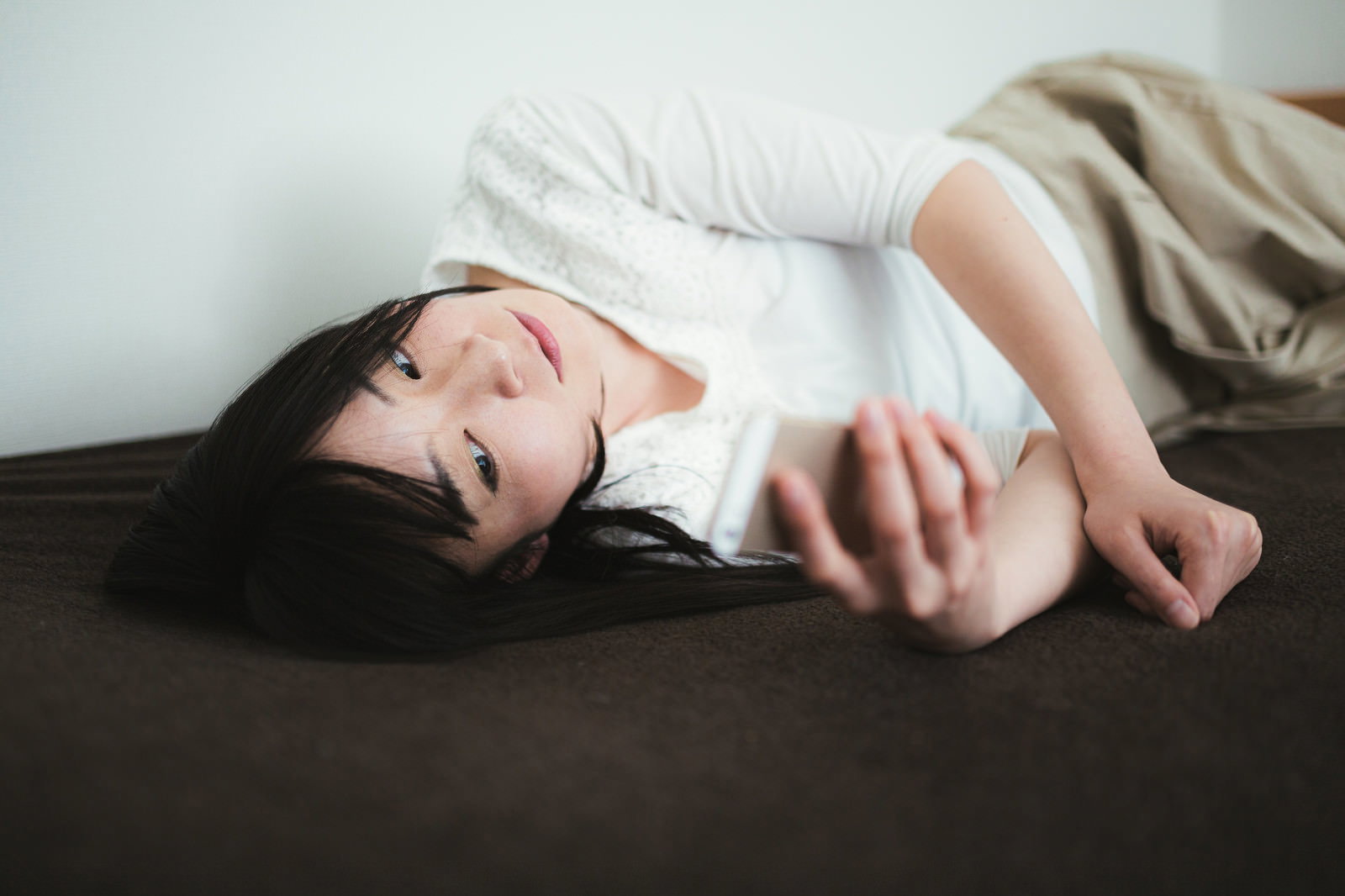 ベッドで横たわりスマートフォンを手に持つ女性が画面を見つめている［モデル：たけべともこ］