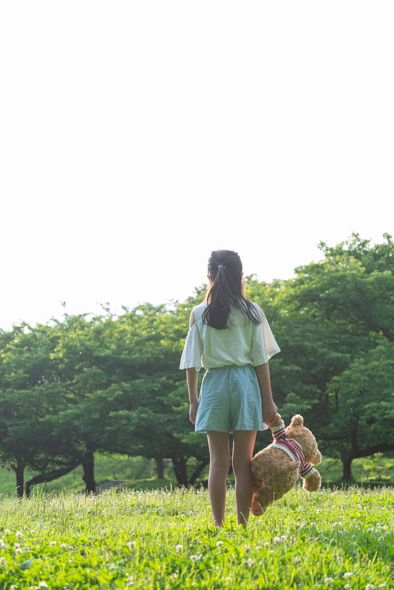 緑の公園でテディベアのぬいぐるみを抱きしめる女の子の後ろ姿