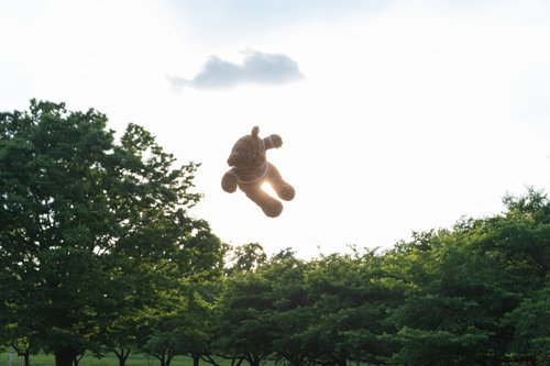 青空と雲を背景に空中を浮かぶクマのぬいぐるみ