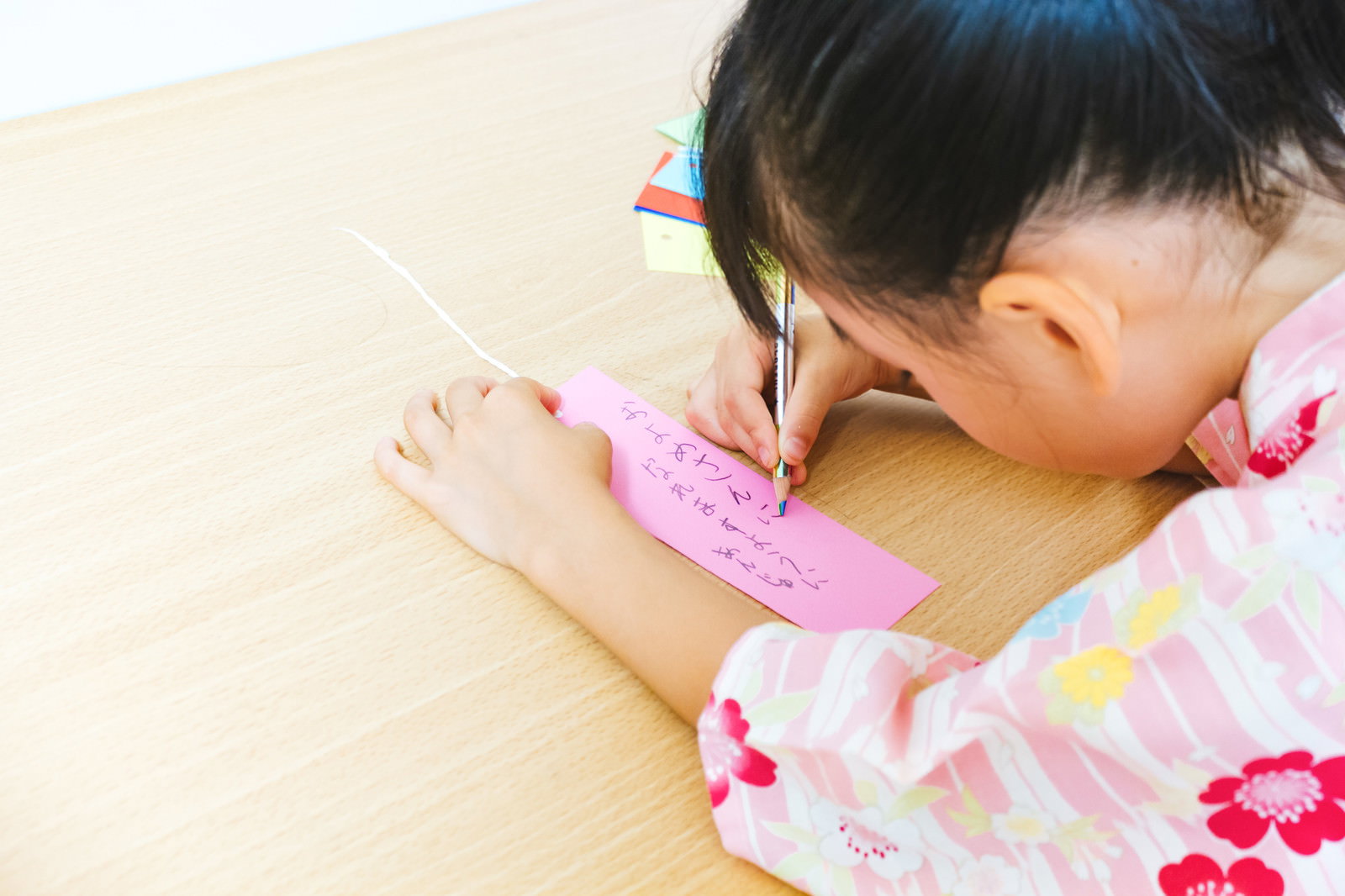 浴衣姿の女の子が床に座り、ピンクの短冊にペンで願い事を書いている［モデル：あんじゅ］