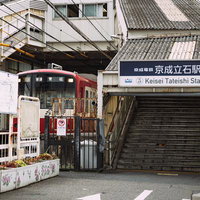 京成立石駅で発車を待つ青砥行の赤い電車と駅舎風景の写真