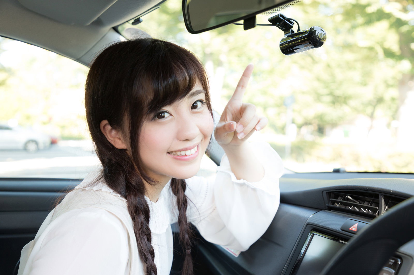 車内でドライブレコーダーを指差す白いトップスの女性［モデル：河村友歌］