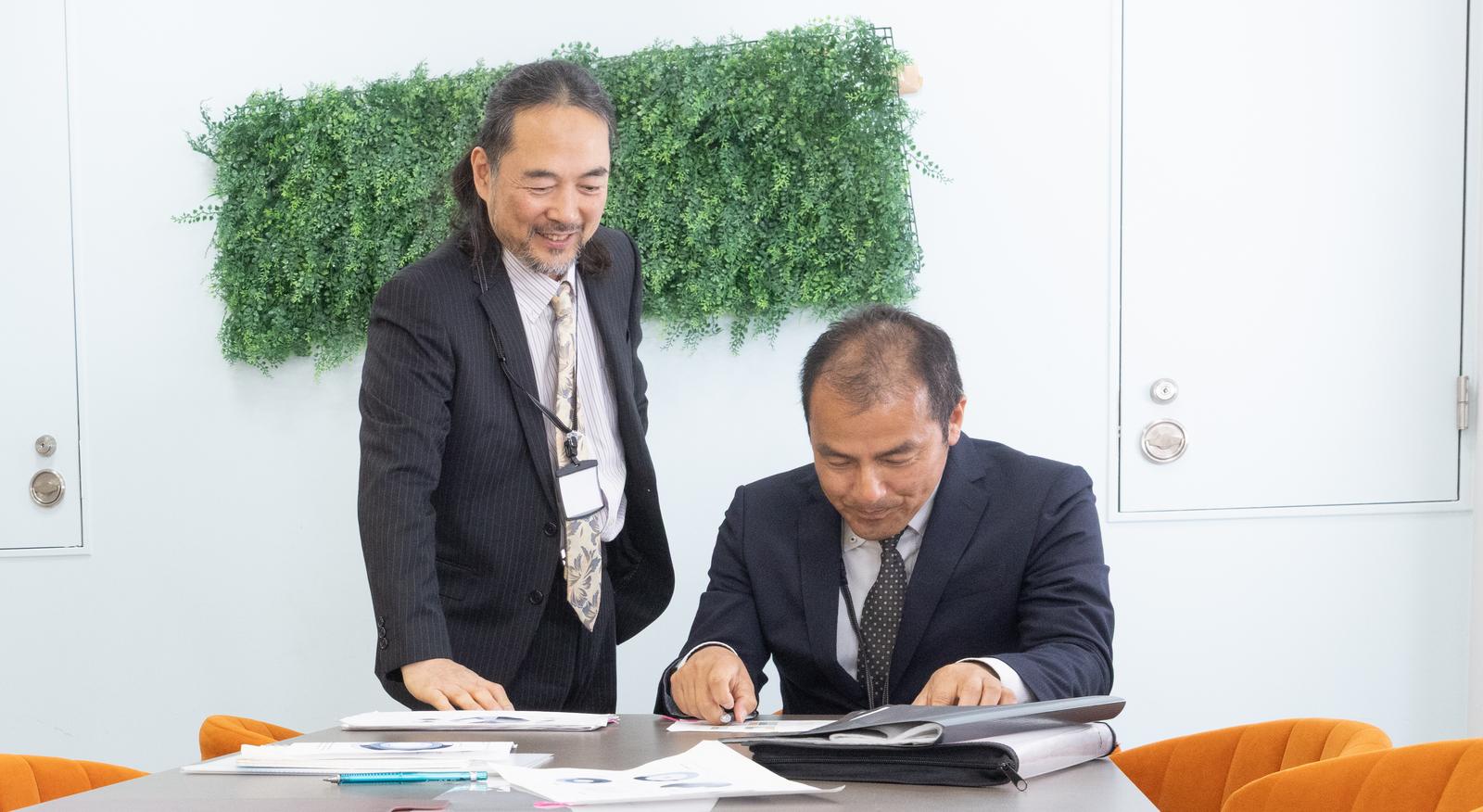 資料を確認する2人の管理職の男性がデスクで向き合う会議シーン［モデル：平野 高木桂一］