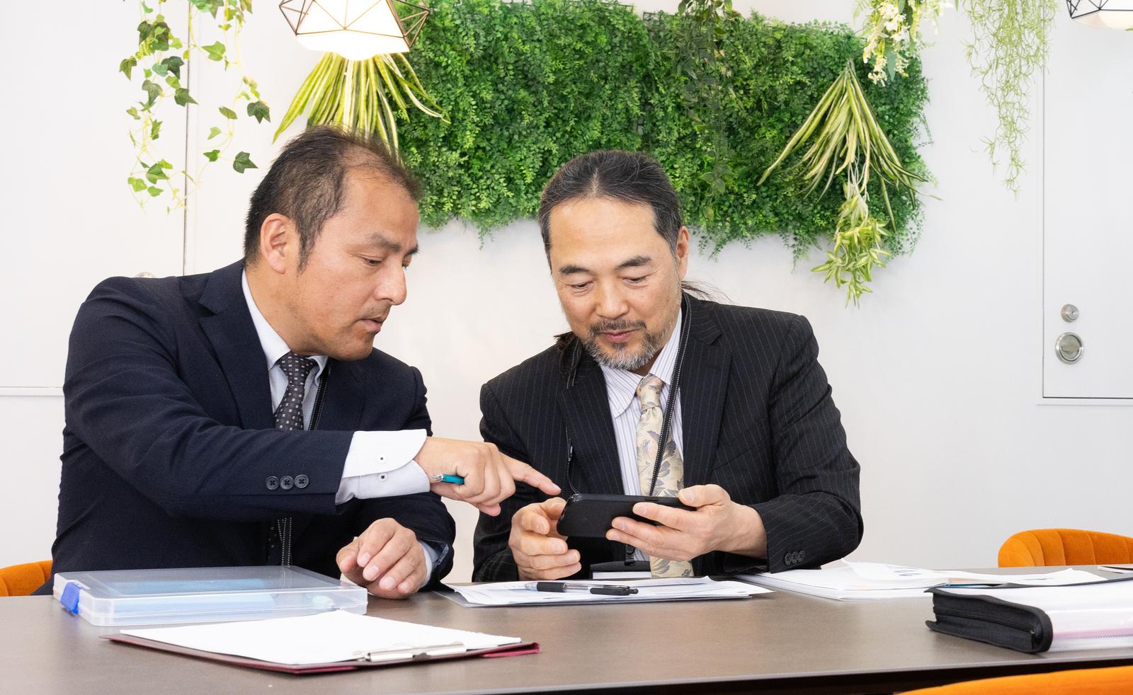 スーツ姿の男性2人がスマホ画面を指さして確認［モデル：高木桂一 平野景由］