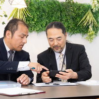 スマホに送られてきたデータを紙の資料で確認する管理職の写真