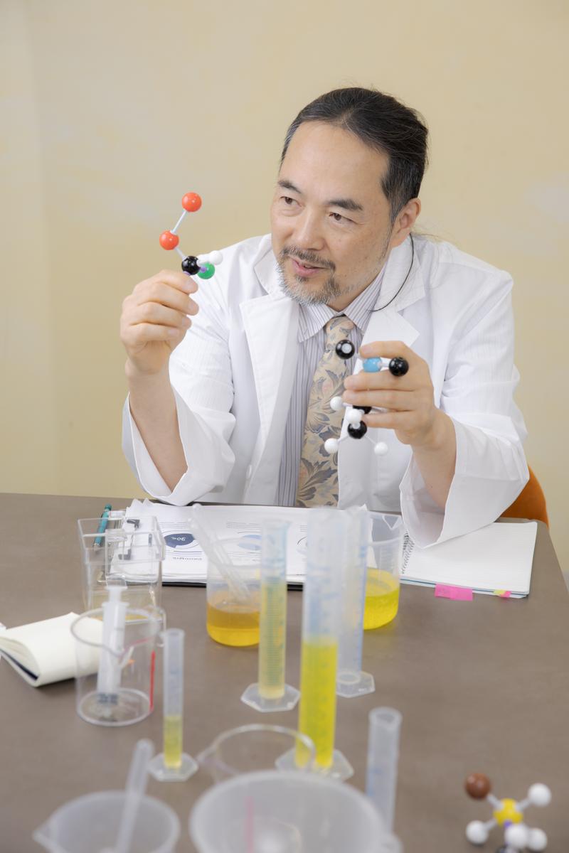 分子モデルを手に持って示す白衣姿の男性研究者［モデル：平野］