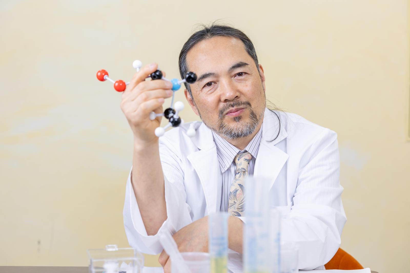 分子モデルを手に持つ白衣姿の男性研究者［モデル：平野］