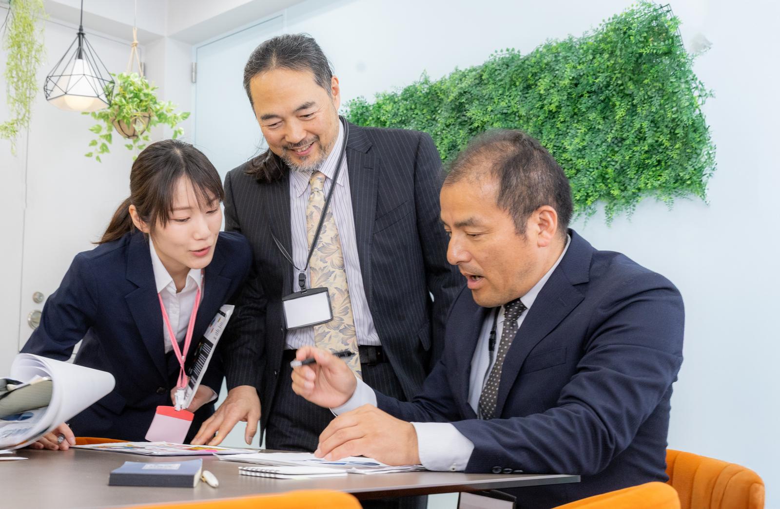 転職したばかりの中途採用者に紙の資料で説明する女性と役員たち［モデル：平野 高木桂一 SAKI］