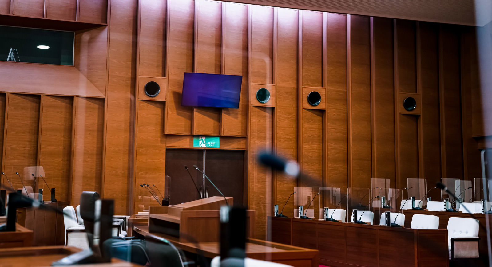 電光板が設置された木製パネルの議場内部、議員席に並ぶ白い椅子と演台