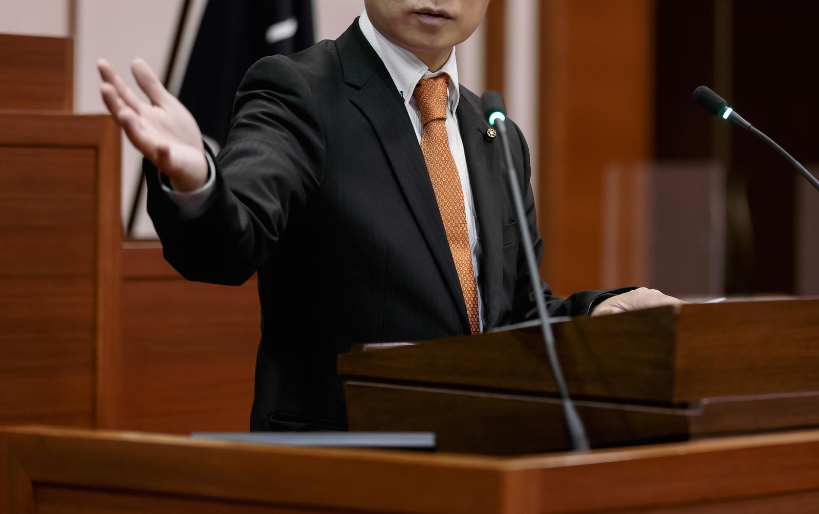 議場で熱心に答弁する黒いスーツの議員と橙色のネクタイ
