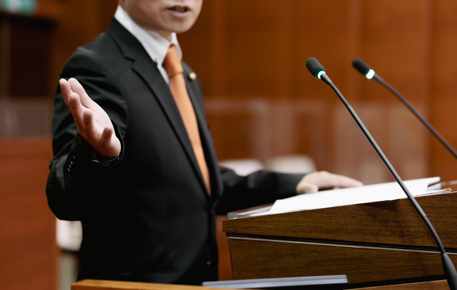 地方議会で黒いスーツの議員が手を広げて激しく追求する様子