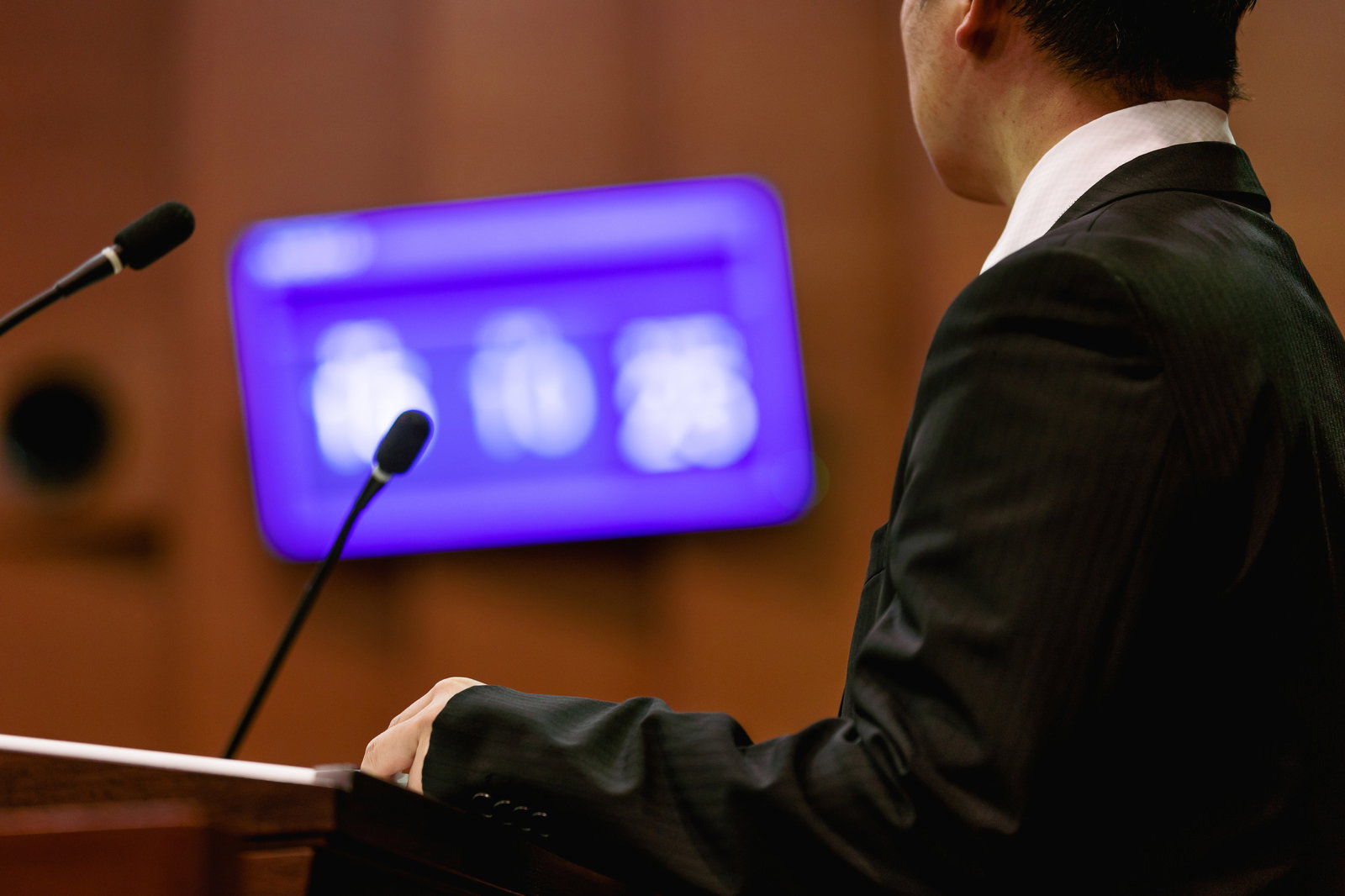 採決の電光板を見つめる市議会議員。スーツ姿で緊張感ある表情。