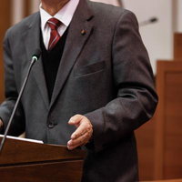 津山市議会でマイクに向かい赤いネクタイの男性が演説する様子の写真