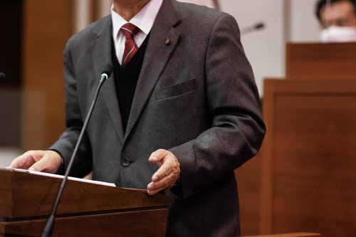 津山市議会でマイクに向かい赤いネクタイの男性が演説する様子