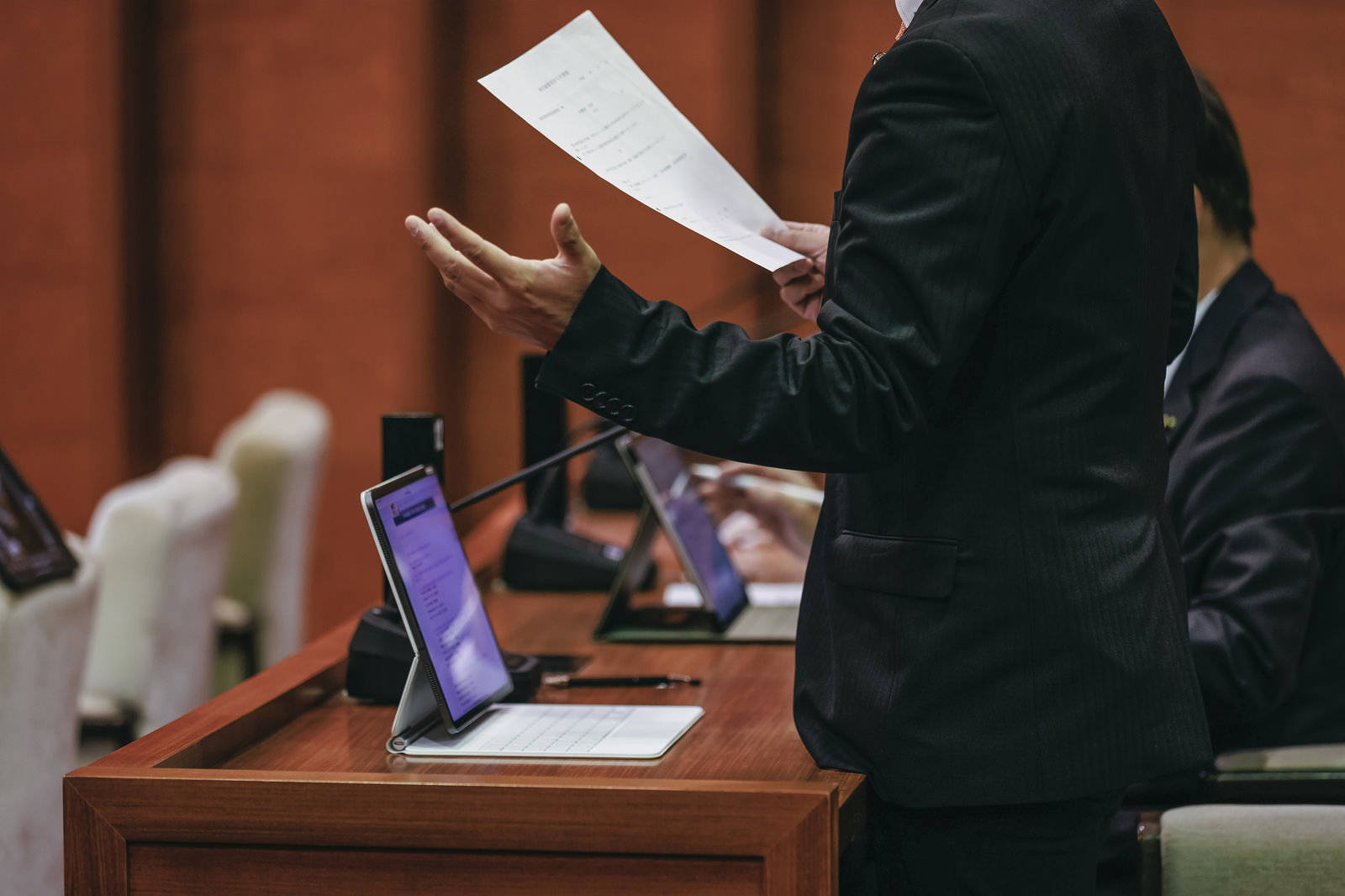 紙の資料を手に議論する市議会議員とタブレット端末が並ぶ議場