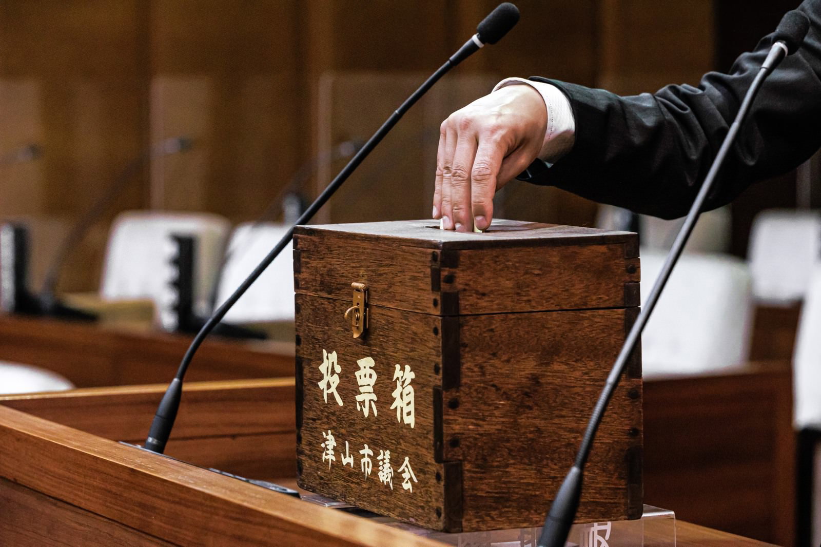 津山市議会の木製投票箱に白い投票用紙を投じる、スーツ姿の男性の手