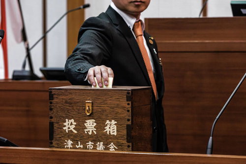 ここまで来て投票に迷いが生じて固まる市議会議員が投票箱に手を伸ばす瞬間