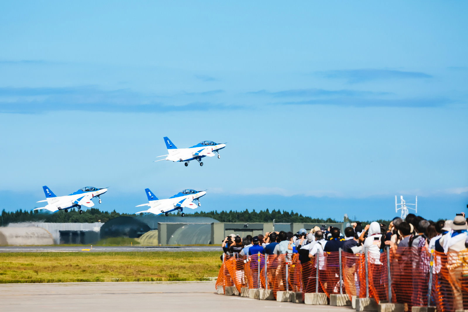 青空の下を飛行するブルーインパルス3機と滑走路脇で見学する観客たち