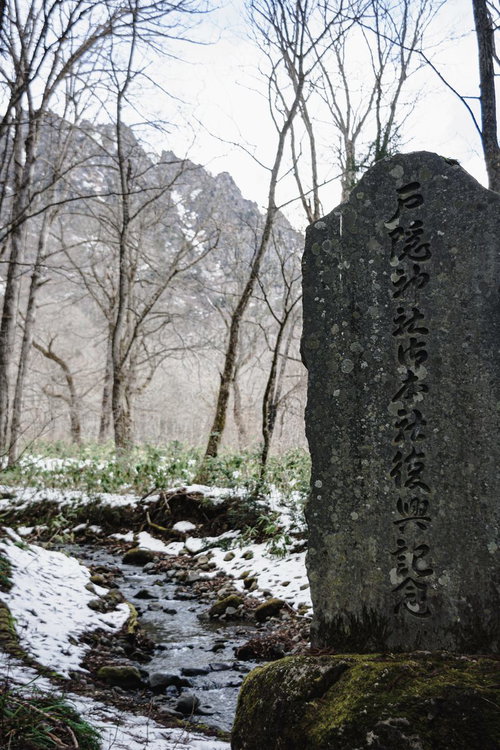 冬枯れた木々の向こうに見える戸隠山や雪の残る逆さ川を背に建つ神社御本社復興記念碑