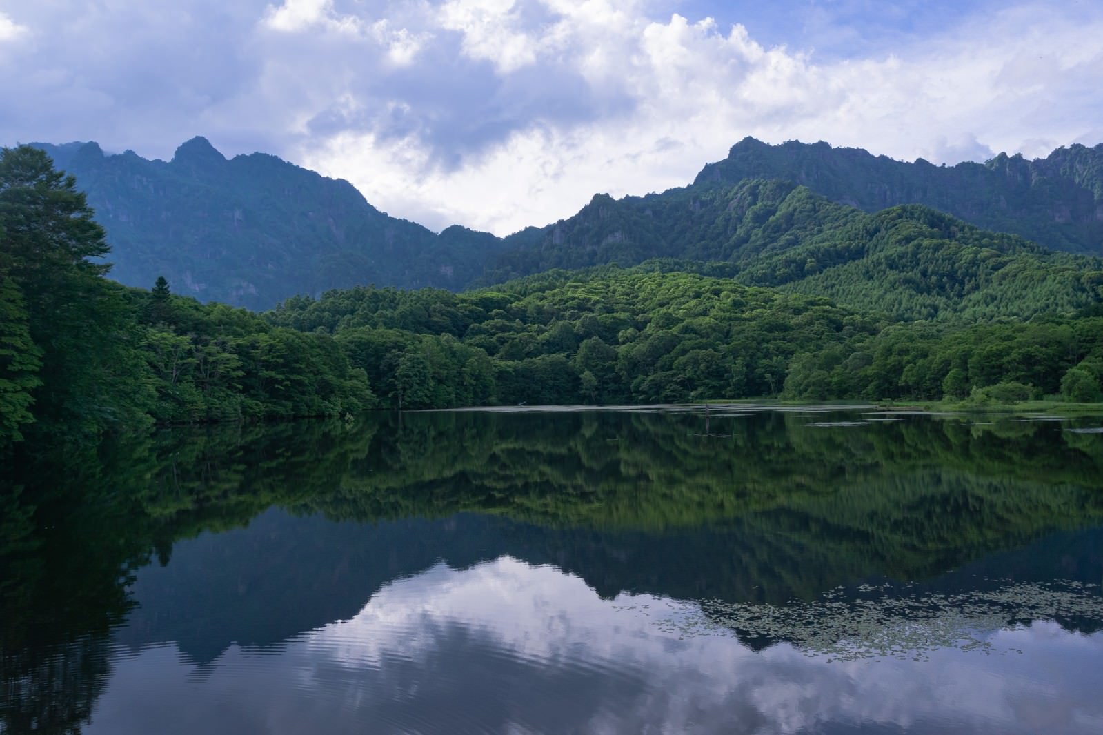 静かな鏡池の水面に映る緑の山並みと森林、青空に浮かぶ雲