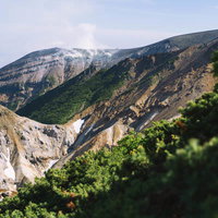 噴煙上げる十勝岳の火口と麓の緑の谷間の自然風景の写真