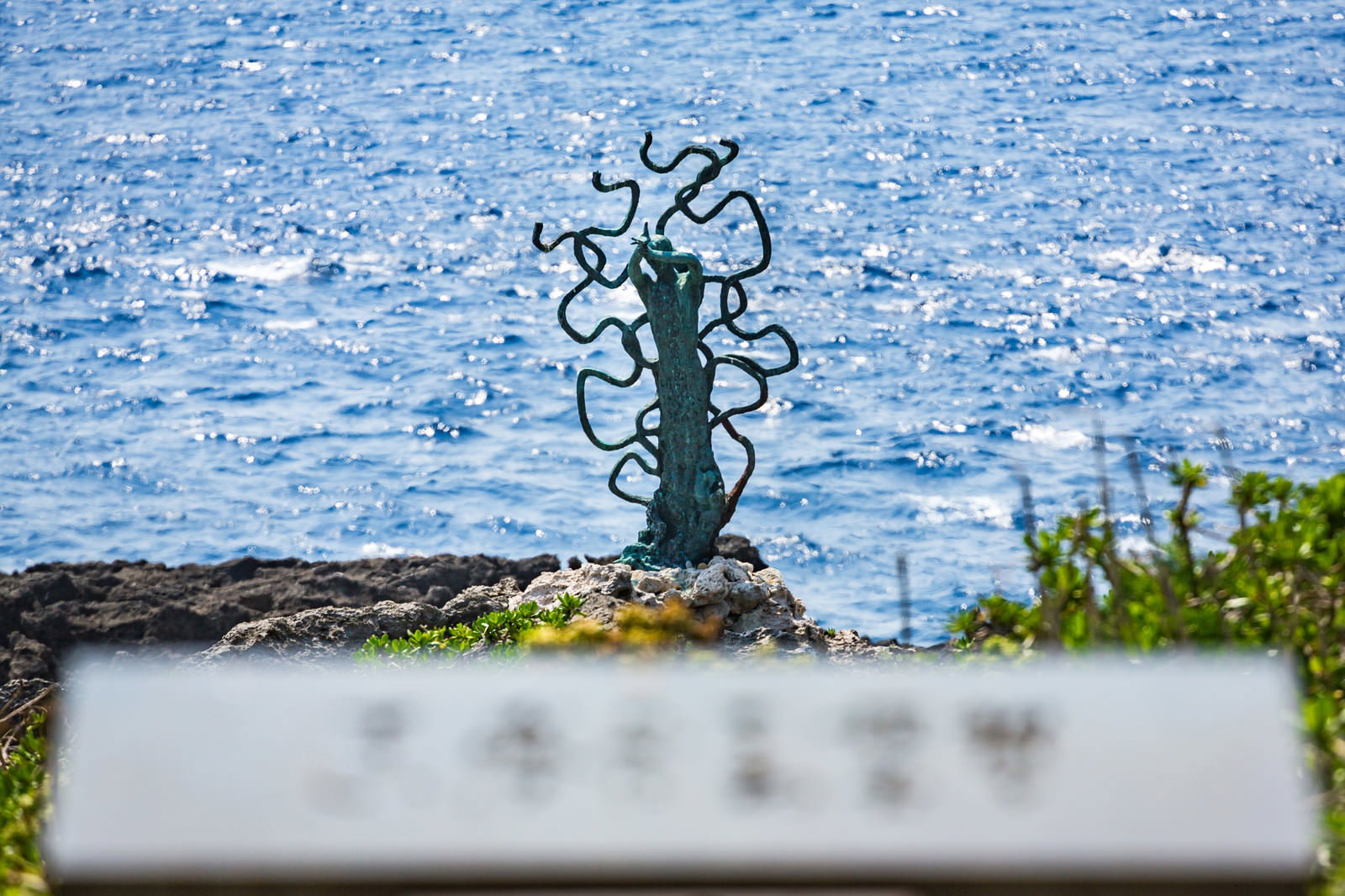 青い海を背景に、岩の上に立つ緑色の銅製の像が両腕を曲げた姿で展示されている